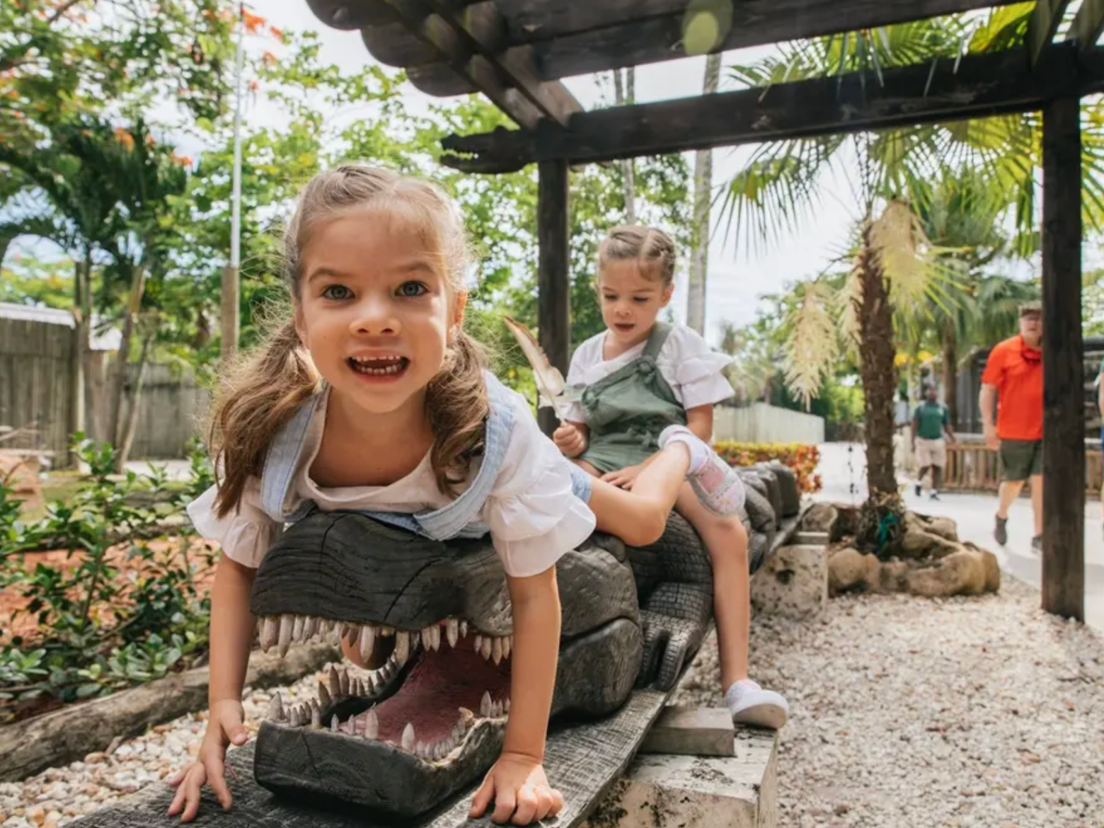 Two children playing on a dinosaur sculpture in a sunny park.